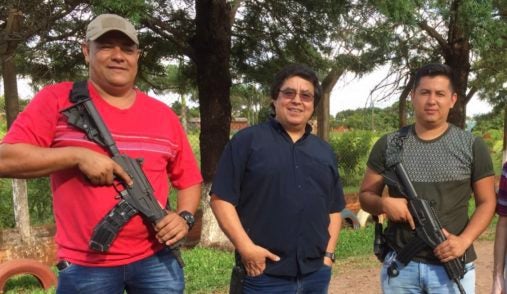 Man stands facing the camera, smiling, with pockets his pants, while two armed individuals stand next to him carrying machine guns to protect him