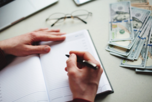 Person writing in a notebook with cash money on the side