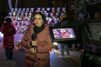 Photo of young woman in a puffy jacket with a microphone in front of photos of people killed in Ukraiine