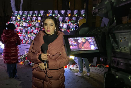 Photo of young woman in a puffy jacket with a microphone in front of photos of people killed in Ukraine