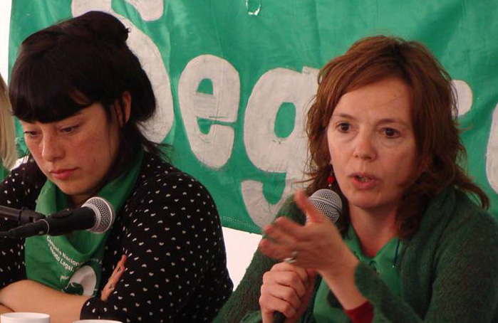 two women wearing green bandanas, one of them speaking on a microphone, with a green background