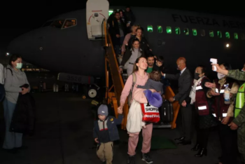 Woman carrying a baby and holding a child's hand at the bottom of the airplane stairway greets reporters.
