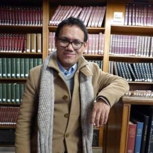 Man with glasses, wearing a wool coat and a scarf looks at the camera. Bookshelves in the background.