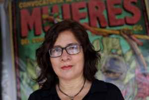 Woman in a black shirt and glasses in front of a wall with a mural and the word "mujeres" (women)