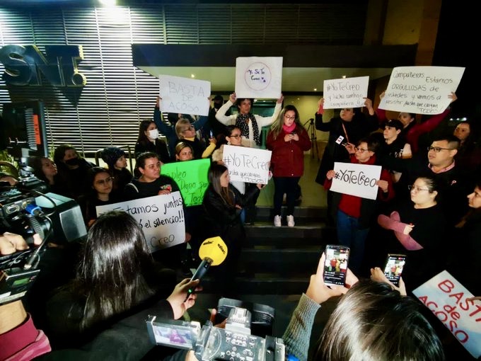 Women in Paraguay holding protest signs, cameras and microphones pointed towards them, night time