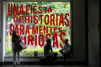 one person standing and two people sitting and chatting in front of a mural written "a party of stories for curious minds" in spanish