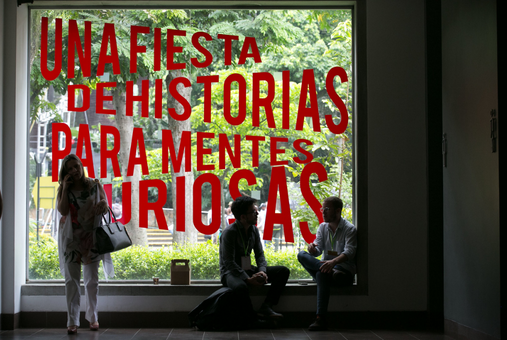 one person standing and two people sitting and chatting in front of a mural written "a party of stories for curious minds" in spanish