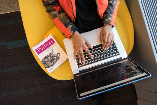 Person typing code on a laptop with a book on Python by their side