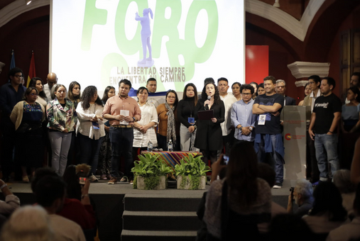 A group of journalists on a stage surrounding a woman speaking on a microphone