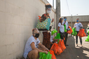 Grupo de pessoas contra uma parede no Brasil, segurando grandes sacolas verdes e laranja e usando máscaras.