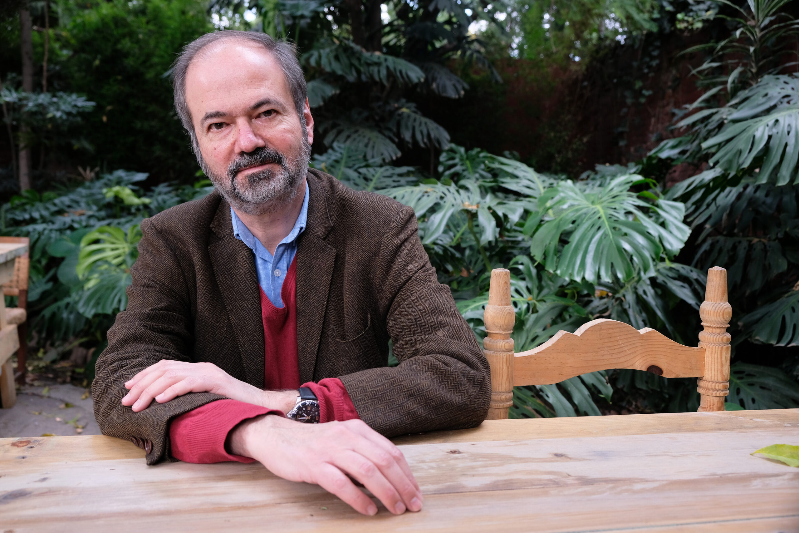 Man with goatee sitting at a wooden table with some plants in the background