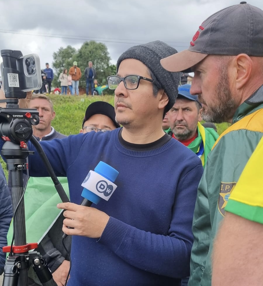 Journalist in blue shirt and beanie is watched over by protesters in Brazil