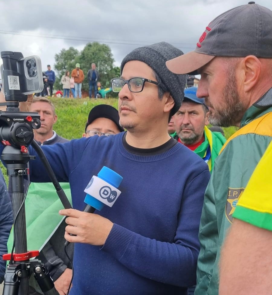 Journalist in blue shirt and beanie is watched over by protesters in Brazil