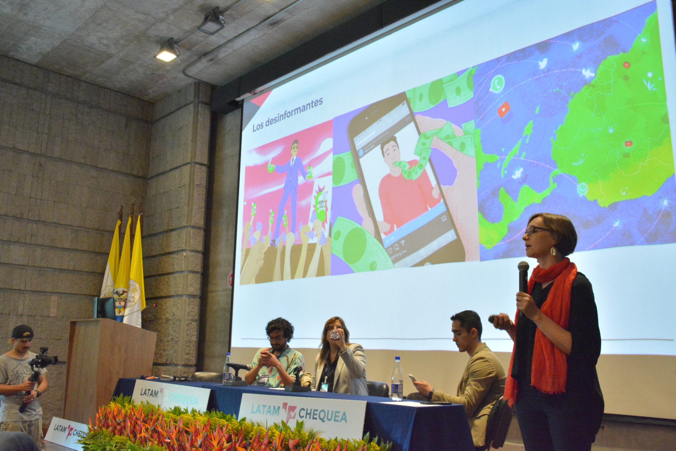 A woman holding a microphone speaks next to a table of panelists and behind is a slide with images illustrating disinformation