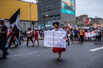 Indigenous woman holding a sign walks on a street in Lima, Peru, with people in the background.