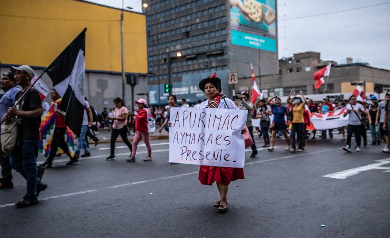 Indigenous woman holding a sign walks on a street in Lima, Peru, with people in the background.