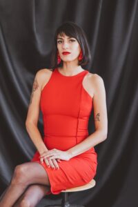 Slim woman with short black hair, red earrings and dress poses in front of the camera, sitting on a stool