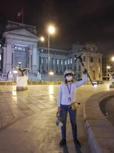 young woman holding a video camera in one hand and a tripod in the other hand stand in front of a government building in Peru, at night