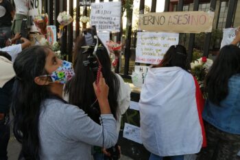 Young woman journalist with long black hair points a camera to a fence covered with protest signs in Peru