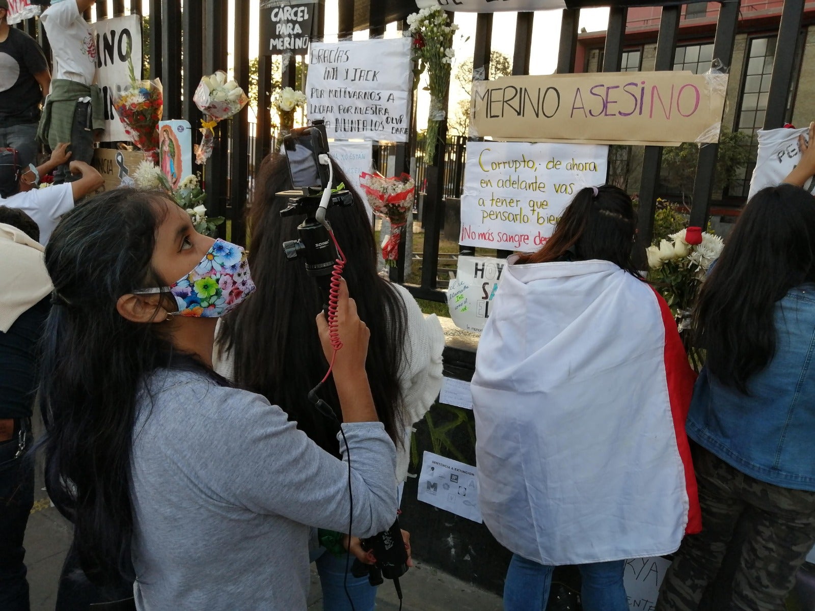 Young woman journalist with long black hair points a camera to a fence covered with protest signs in Peru