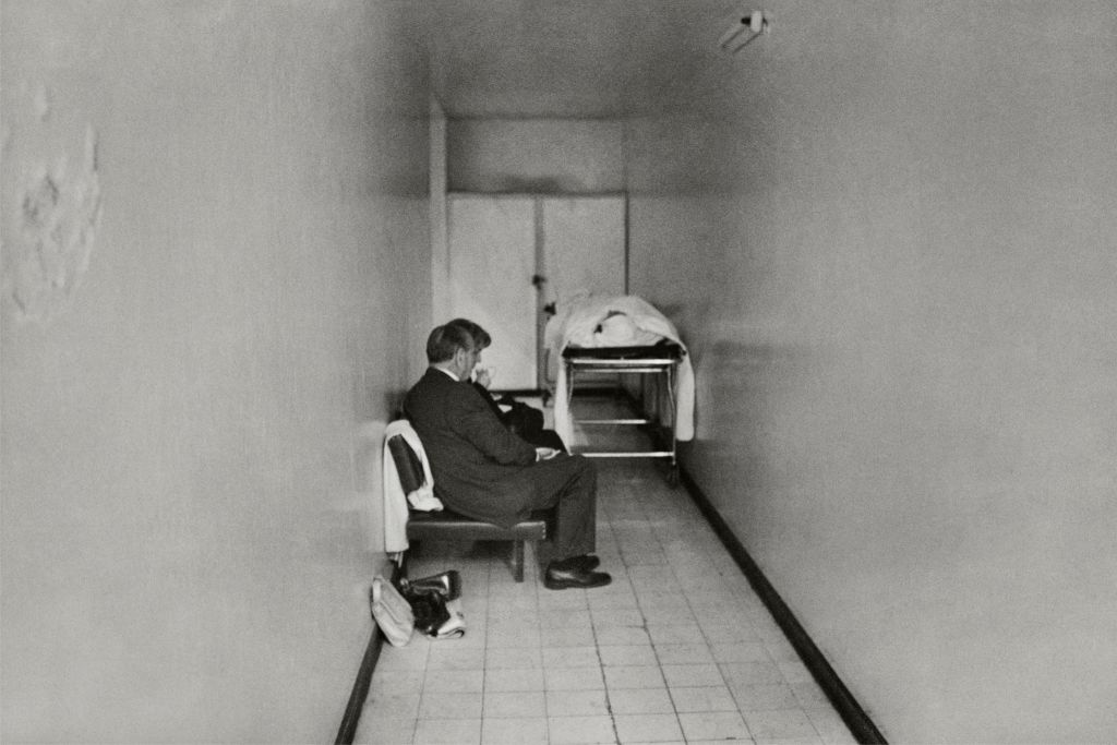 Pasillo de un hospital con una camilla con el cuerpo de Pablo Neruda tapado por una sábana en la esquina superior derecha mientras un hombre y una mujer están sentados en frente