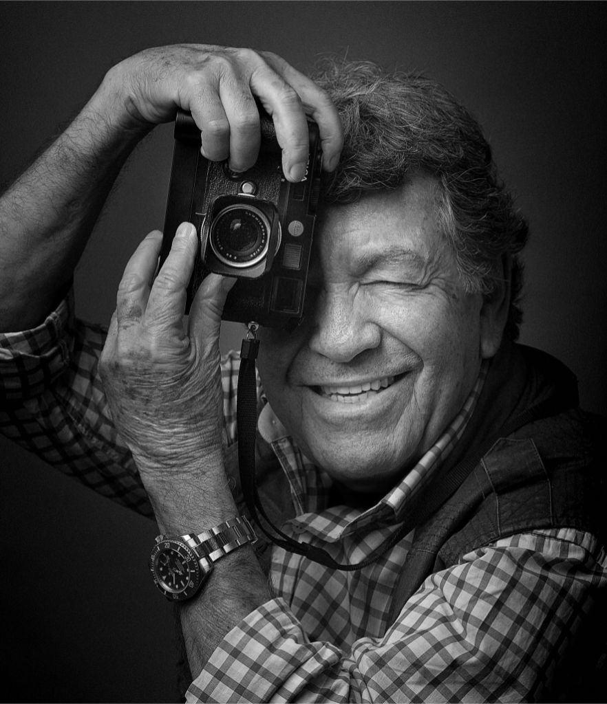 Retrato en blanco y negro de un hombre mayor sosteniendo una cámara fotográfica como si estuviera tomando una foto a su observador
