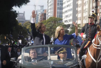 A man in a suit and a woman in a blue dress wave to the crowds when being inaugurated as president and vice-president of Uruguay. A horse and soldier to the right and a man follows along on foot to the left.