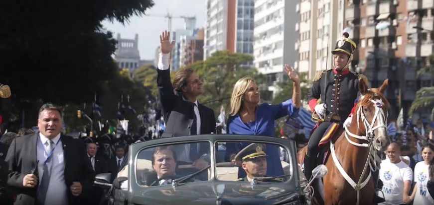 A man in a suit and a woman in a blue dress wave to the crowds when being inaugurated as president and vice-president of Uruguay. A horse and soldier to the right and a man follows along on foot to the left.