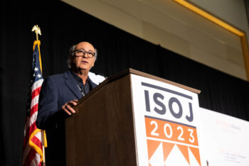 A man with glasses stands at a podium with a United States flag to the left and the ISOJ 2023 sign on podium