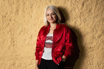 Woman with platinum blonde hair in a shiny red jacket posing in front of a wall