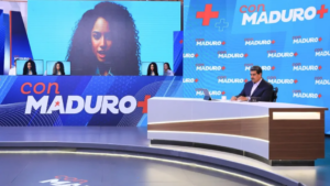 Set te un programa de televisión. En la mesa principal está sentado Nicolás Maduro con traje negro, atrás y al lado está una pantalla donde se ve la presentadora creada con inteligencia artificial.