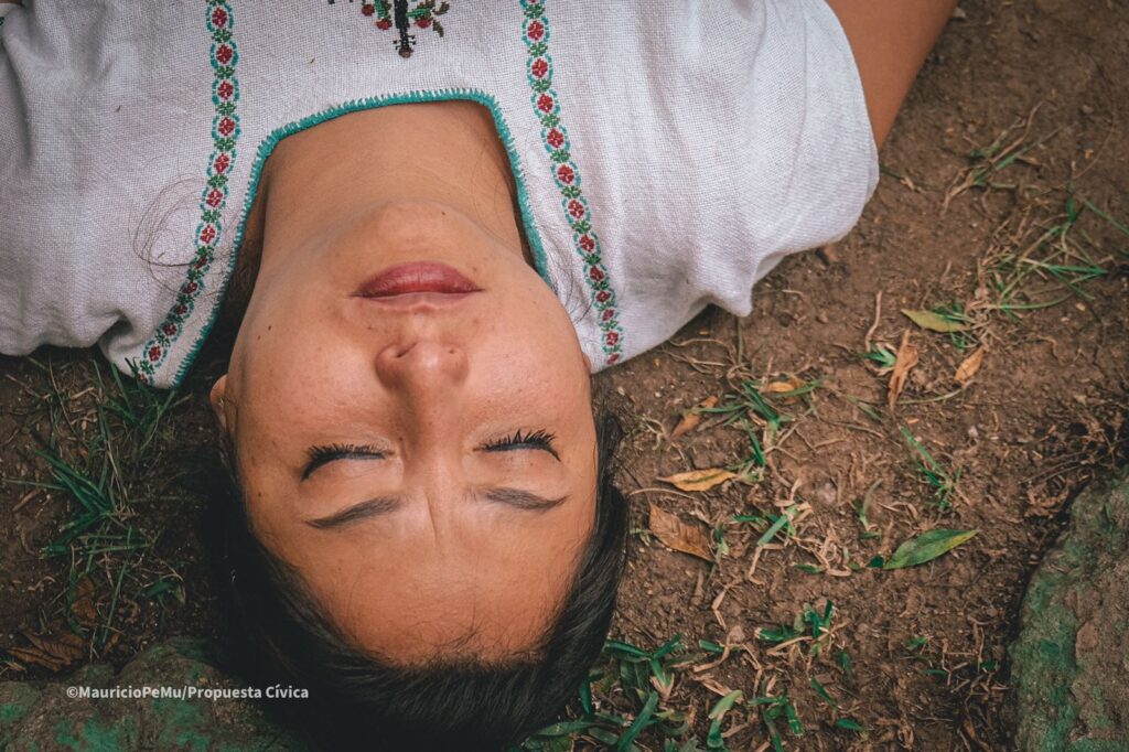 Fotografía de una mujer acostada en un suelo de tierra y prado, con los ojos cerrados y de cabeza. 