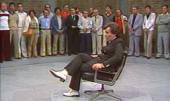 Hombre con traje negro y zapatos blancos sentado en una silla en el centro de un set de televisión rodeado de personas
