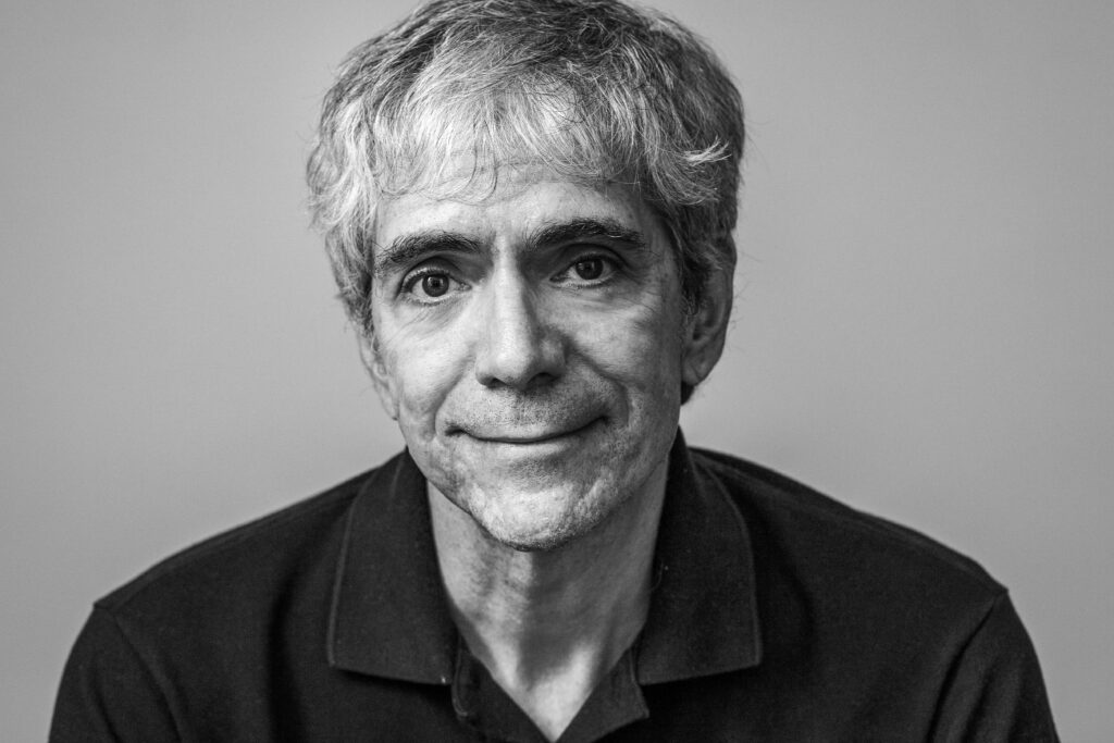 Fotografía en blanco y negro de un hombre hasta sus hombros con camisa oscura, sonriendo a la cámara