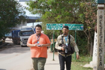 Two men, one with a notebook and the other one with a camera, walk towards the camera at a border crossing in South America