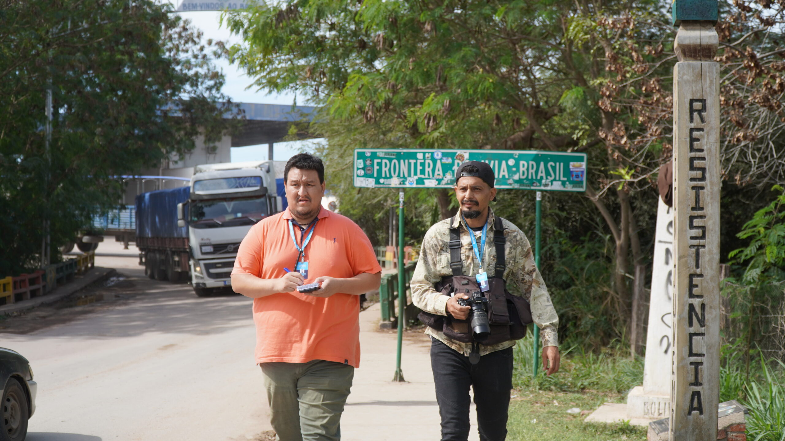 Two men, one with a notebook and the other one with a camera, walk towards the camera at a border crossing in South America