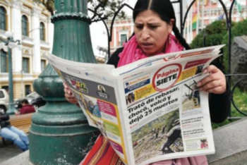 A woman in the middle of a street plaza reads a newspaper while sitting in a bench
