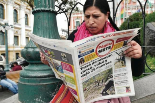 A woman in the middle of a street plaza reads a newspaper while sitting in a bench