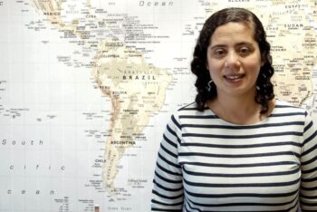 Woman with curly hair and a striped black and white top stands in front of a map featuring South America