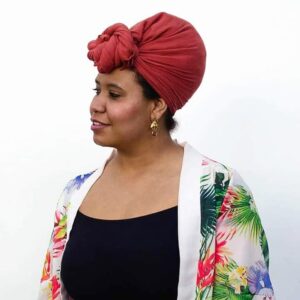 Black woman with a red kerchief on her head and a flowered top over a black tank top looks sideway, smiling
