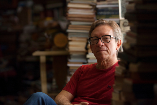 man looking at the camera and sitting amidst piles of books