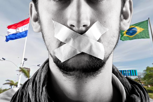 man with some tape on his mouth cannot speak, with the flags of Brazil and Paraguay in the background