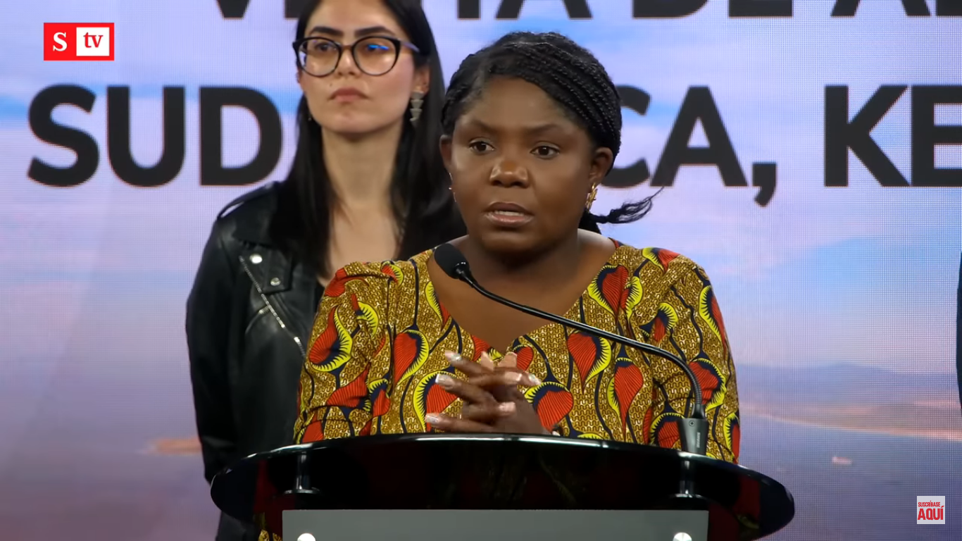 Black woman with colorful red, yellow and black dress speaks before a microphone while another woman with glasses watches from behind