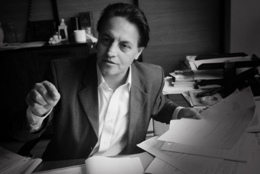 Black-and-white photo of a man with dark hair seated at a table cluttered with papers. He is holding a page in his left hand and gesturing with his right hand as if speaking or making a point