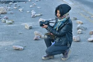 woman with leather jacket and scarf adjusts the camera while crouching on the street surrounded by rocks
