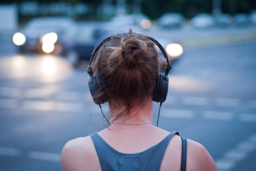 A woman immersed in a story, wearing headphones while standing on a bustling street with cars in the foreground