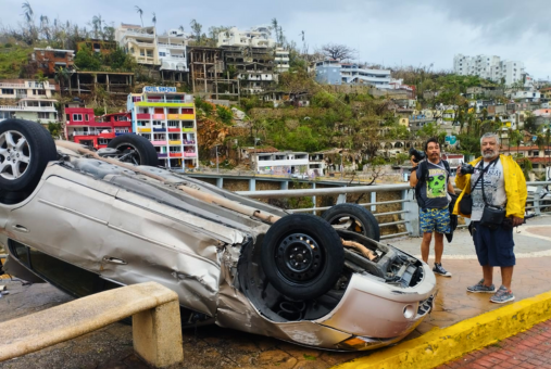 Two photojournalists hold their cameras pose next to a car overturned by Hurricane Otis strong winds.