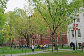 Grupo de alunos caminha em um gramado sob árvores em clima outonal. À direita, é possível ver o prédio de tijolos de um prédio de Harvard