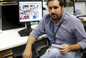 Um retrato do jornalista Rafael Soares sentado em sua mesa na redação dos jornais O Globo e Extra, no Rio de Janeiro. Ele tem barba e cabelo castanho, é moreno e está segurando um bloco de papel enquanto encara a câmera