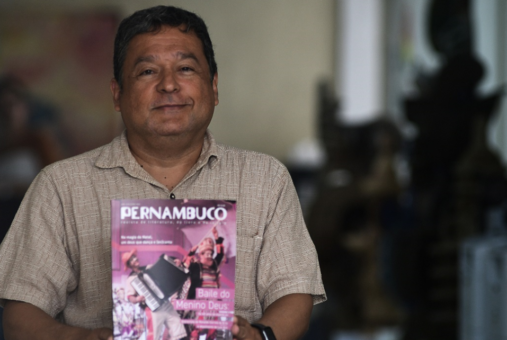 A portrait of a man wearing a brown shirt and holding a copy a Pernambuco magazine